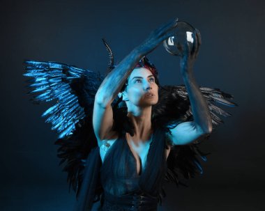 Gotik boynuzlu başlık giyen kadın modelin yakın portresi, cadılar bayramı siyah elbisesi, fantezi melek tüyü kanatları. Mistik kristal küreyi tutuyorum. Karanlık stüdyo arka planı, sinematik gölge silueti 
