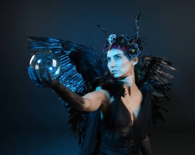 Gotik boynuzlu başlık giyen kadın modelin yakın portresi, cadılar bayramı siyah elbisesi, fantezi melek tüyü kanatları. Mistik kristal küreyi tutuyorum. Karanlık stüdyo arka planı, sinematik gölge silueti 