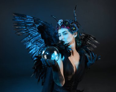 Gotik boynuzlu başlık giyen kadın modelin yakın portresi, cadılar bayramı siyah elbisesi, fantezi melek tüyü kanatları. Mistik kristal küreyi tutuyorum. Karanlık stüdyo arka planı, sinematik gölge silueti 