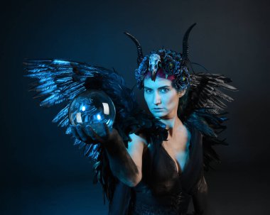 Gotik boynuzlu başlık giyen kadın modelin yakın portresi, cadılar bayramı siyah elbisesi, fantezi melek tüyü kanatları. Mistik kristal küreyi tutuyorum. Karanlık stüdyo arka planı, sinematik gölge silueti 
