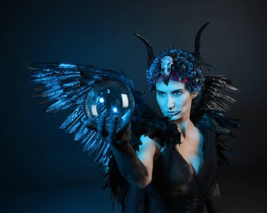 Gotik boynuzlu başlık giyen kadın modelin yakın portresi, cadılar bayramı siyah elbisesi, fantezi melek tüyü kanatları. Mistik kristal küreyi tutuyorum. Karanlık stüdyo arka planı, sinematik gölge silueti 