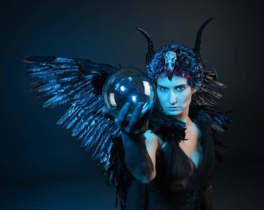 Gotik boynuzlu başlık giyen kadın modelin yakın portresi, cadılar bayramı siyah elbisesi, fantezi melek tüyü kanatları. Mistik kristal küreyi tutuyorum. Karanlık stüdyo arka planı, sinematik gölge silueti 