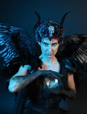 Gotik boynuzlu başlık giyen kadın modelin yakın portresi, cadılar bayramı siyah elbisesi, fantezi melek tüyü kanatları. Mistik kristal küreyi tutuyorum. Karanlık stüdyo arka planı, sinematik gölge silueti 