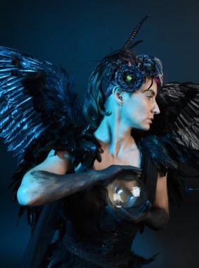Gotik boynuzlu başlık giyen kadın modelin yakın portresi, cadılar bayramı siyah elbisesi, fantezi melek tüyü kanatları. Mistik kristal küreyi tutuyorum. Karanlık stüdyo arka planı, sinematik gölge silueti 