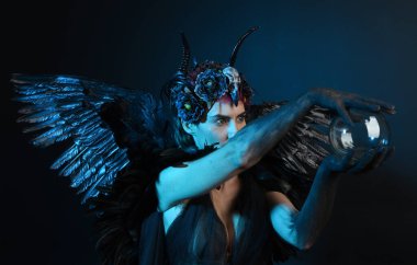 Gotik boynuzlu başlık giyen kadın modelin yakın portresi, cadılar bayramı siyah elbisesi, fantezi melek tüyü kanatları. Mistik kristal küreyi tutuyorum. Karanlık stüdyo arka planı, sinematik gölge silueti 