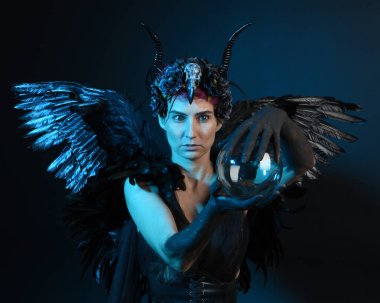 Gotik boynuzlu başlık giyen kadın modelin yakın portresi, cadılar bayramı siyah elbisesi, fantezi melek tüyü kanatları. Mistik kristal küreyi tutuyorum. Karanlık stüdyo arka planı, sinematik gölge silueti 