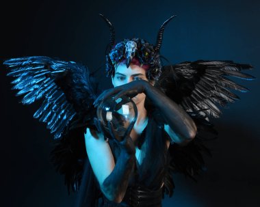 Gotik boynuzlu başlık giyen kadın modelin yakın portresi, cadılar bayramı siyah elbisesi, fantezi melek tüyü kanatları. Mistik kristal küreyi tutuyorum. Karanlık stüdyo arka planı, sinematik gölge silueti 