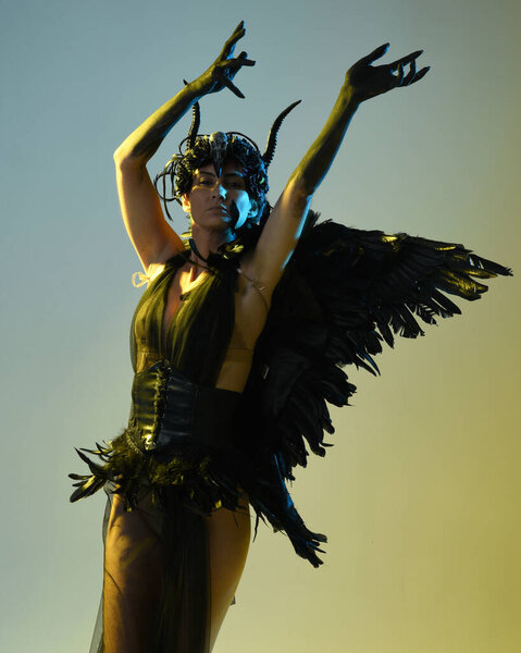 Close up portrait of female model wearing gothic horned headdress, halloween black dress and fantasy angel feather wings. Isolated dark studio background with cinematic shadow silhouettes 