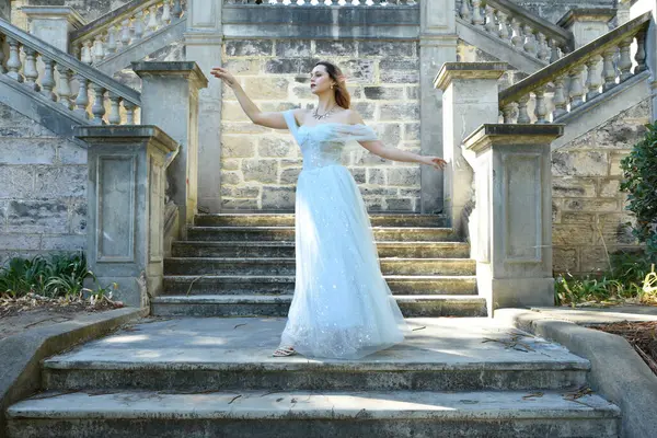 portrait of beautiful female model wearing blue fantasy ballgown, like a fairytale elf princess. standing on staircase, walking up stairs of a romantic castle balcony location.