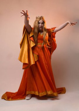 Uzun sarı saçlı, orta çağ fantezi elbisesi giyen, uzun kollu, kraliyet kraliçesinin altın tacını takan uzun boylu bir kadının tam boy portresi. Stüdyo arka planında izole edilmiş, el kol hareketleriyle poz vermek.