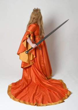 Orta çağ fantezi elbisesi giyen iri yarı sarışın bir kadının tam boy portresi. Altın taç kraliyet kraliçesi. Arka planda durmak, kılıç tutmak, izole edilmiş stüdyo geçmişi..