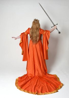 Orta çağ fantezi elbisesi giyen iri yarı sarışın bir kadının tam boy portresi. Altın taç kraliyet kraliçesi. Arka planda durmak, kılıç tutmak, izole edilmiş stüdyo geçmişi..