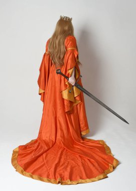 Orta çağ fantezi elbisesi giyen iri yarı sarışın bir kadının tam boy portresi. Altın taç kraliyet kraliçesi. Arka planda durmak, kılıç tutmak, izole edilmiş stüdyo geçmişi..