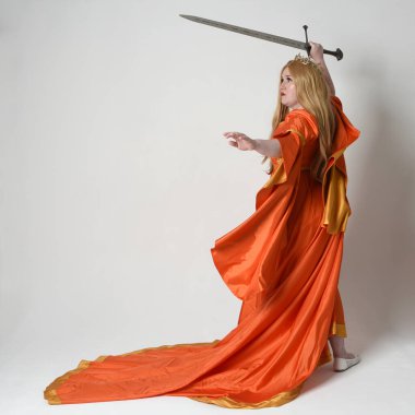 Orta çağ fantezi elbisesi giyen iri yarı sarışın bir kadının tam boy portresi. Altın taç kraliyet kraliçesi. Arka planda durmak, kılıç tutmak, izole edilmiş stüdyo geçmişi..