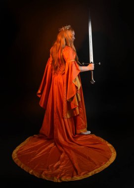 Ortaçağ fantezi elbisesi giyen, kraliyet kraliçesi tacını giymiş, iri yarı sarışın bir kadının tam boy portresi. Arka plan duruşu, kılıç silahı, izole edilmiş karanlık stüdyo arka plan silueti.