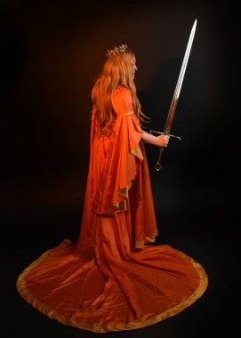 Ortaçağ fantezi elbisesi giyen, kraliyet kraliçesi tacını giymiş, iri yarı sarışın bir kadının tam boy portresi. Arka plan duruşu, kılıç silahı, izole edilmiş karanlık stüdyo arka plan silueti.