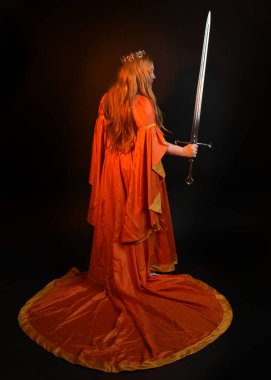 Ortaçağ fantezi elbisesi giyen, kraliyet kraliçesi tacını giymiş, iri yarı sarışın bir kadının tam boy portresi. Arka plan duruşu, kılıç silahı, izole edilmiş karanlık stüdyo arka plan silueti.