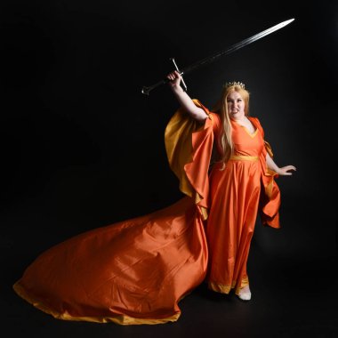 Ortaçağ fantezi elbisesi giyen, kraliyet kraliçesi tacını giymiş, iri yarı sarışın bir kadının tam boy portresi. Arka plan duruşu, kılıç silahı, izole edilmiş karanlık stüdyo arka plan silueti.