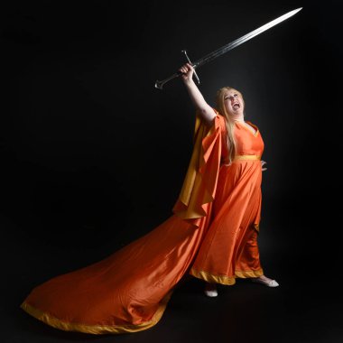 Ortaçağ fantezi elbisesi giyen, kraliyet kraliçesi tacını giymiş, iri yarı sarışın bir kadının tam boy portresi. Arka plan duruşu, kılıç silahı, izole edilmiş karanlık stüdyo arka plan silueti.