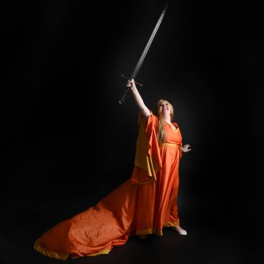 Ortaçağ fantezi elbisesi giyen, kraliyet kraliçesi tacını giymiş, iri yarı sarışın bir kadının tam boy portresi. Arka plan duruşu, kılıç silahı, izole edilmiş karanlık stüdyo arka plan silueti.