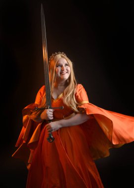 Ortaçağ fantezi elbisesi giyen, kraliyet kraliçesi tacını giymiş, iri yarı sarışın bir kadının portresi. Arka plan duruşu, kılıç silahı, izole edilmiş karanlık stüdyo arka plan silueti.