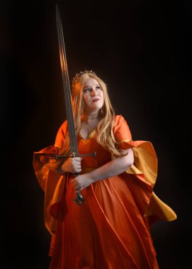 Ortaçağ fantezi elbisesi giyen, kraliyet kraliçesi tacını giymiş, iri yarı sarışın bir kadının portresi. Arka plan duruşu, kılıç silahı, izole edilmiş karanlık stüdyo arka plan silueti.