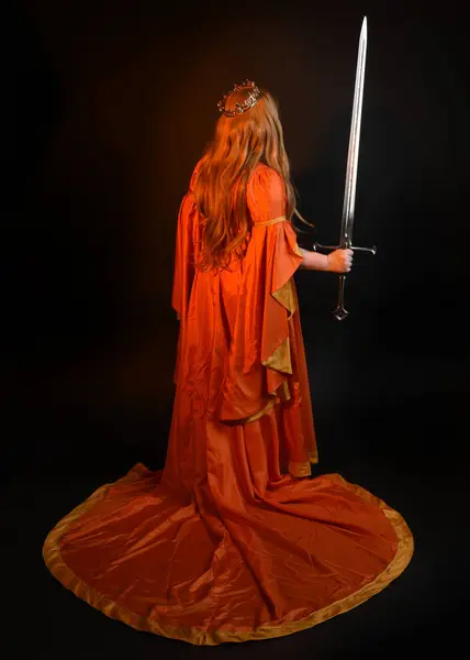 Full length portrait of plus size blonde woman wearing historical medieval fantasy gown, crowned royal queen. Standing pose backview, holding sword weapon, isolated dark studio background silhouette