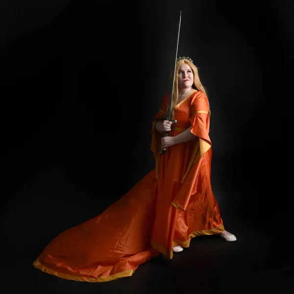 Full length portrait of plus size blonde woman wearing historical medieval fantasy gown, crowned royal queen. Standing pose backview, holding sword weapon, isolated dark studio background silhouette