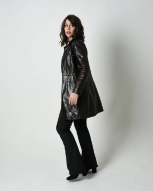 Uzun deri yağmurluk giyen esmer kadının tam boy portresi. Kameradan uzaklaşarak poz vermek, arkana bakmak, uzanmak. Beyaz stüdyo arka planında izole edilmiş..