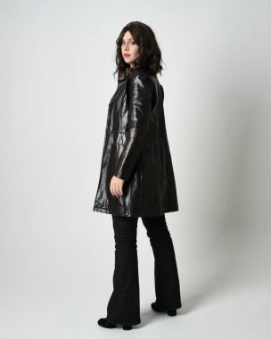 Uzun deri yağmurluk giyen esmer kadının tam boy portresi. Kameradan uzaklaşarak poz vermek, arkana bakmak, uzanmak. Beyaz stüdyo arka planında izole edilmiş..