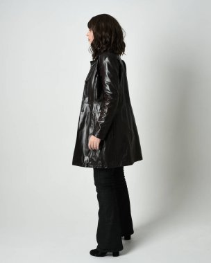 Uzun deri yağmurluk giyen esmer kadının tam boy portresi. Yan profilde duruyor, omzunun üstünden geriye bakıyor, uzanıyor. Beyaz stüdyo arka planında izole edilmiş..