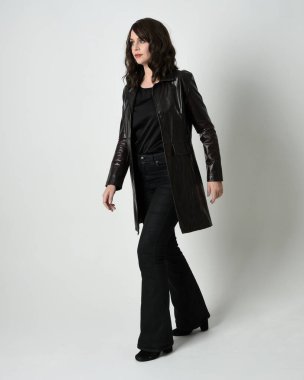 Uzun deri yağmurluk giyen esmer kadının tam boy portresi. Yan profilde duruyor, omzunun üstünden geriye bakıyor, uzanıyor. Beyaz stüdyo arka planında izole edilmiş..