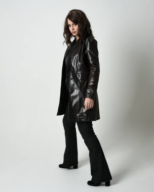 Uzun deri yağmurluk giyen esmer kadının tam boy portresi. Yan profilde duruyor, omzunun üstünden geriye bakıyor, uzanıyor. Beyaz stüdyo arka planında izole edilmiş..