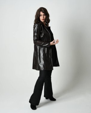 Uzun deri yağmurluk giyen esmer kadının tam boy portresi. Yan profilde duruyor, omzunun üstünden geriye bakıyor, uzanıyor. Beyaz stüdyo arka planında izole edilmiş..