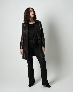 Uzun siyah deri trençkot giyen güzel esmer kadının tam boy portresi, kara film dedektifi. Poz vermek, kameraya doğru yürümek, uzanmak. İzole edilmiş beyaz stüdyo arka planı