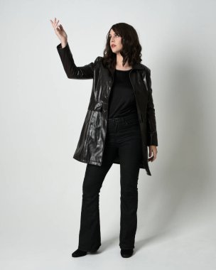 Uzun siyah deri trençkot giyen güzel esmer kadının tam boy portresi, kara film dedektifi. Poz vermek, kameraya doğru yürümek, uzanmak. İzole edilmiş beyaz stüdyo arka planı