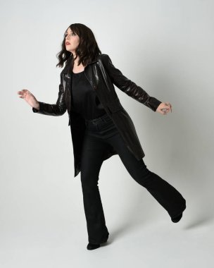 Uzun siyah deri trençkot giyen güzel esmer kadının tam boy portresi, kara film dedektifi. Poz vermek, kameraya doğru yürümek, uzanmak. İzole edilmiş beyaz stüdyo arka planı