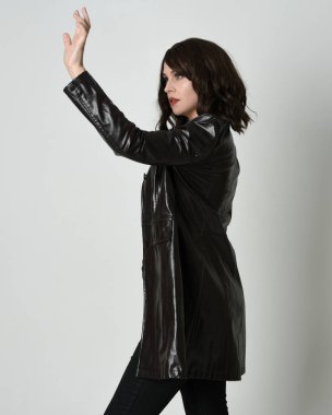 Uzun siyah deri trençkot giyen güzel esmer kadının portresi, kara film dedektifi. Coğrafi el pozları, kollar uzanmış, stüdyo arka planında izole edilmiş.