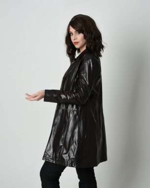 Uzun siyah deri trençkot giyen güzel esmer kadının portresi, kara film dedektifi. Coğrafi el pozları, kollar uzanmış, stüdyo arka planında izole edilmiş.