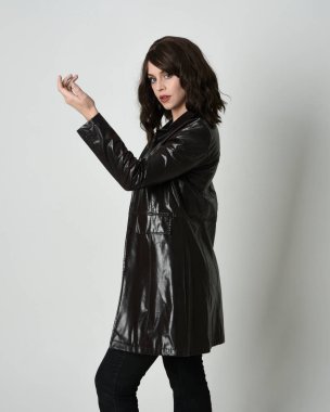 Uzun siyah deri trençkot giyen güzel esmer kadının portresi, kara film dedektifi. Coğrafi el pozları, kollar uzanmış, stüdyo arka planında izole edilmiş.