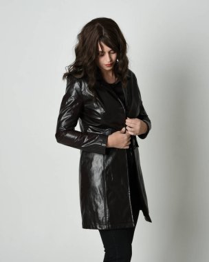 Uzun siyah deri trençkot giyen güzel esmer kadının portresi, kara film dedektifi. Coğrafi el pozları, kollar uzanmış, stüdyo arka planında izole edilmiş.