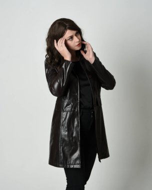 Uzun siyah deri trençkot giyen güzel esmer kadının portresi, kara film dedektifi. Coğrafi el pozları, kollar uzanmış, stüdyo arka planında izole edilmiş.