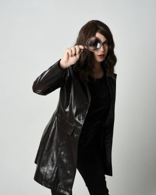 Siyah deri trençkot giyen güzel esmer kadının portresi. Dedektifin büyüteçini tutuyor, kameraya uzanıyor ve bir şey arıyor. İzole stüdyo geçmişi