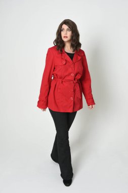 Güzel esmer kadın modelin tam boy portresi, kırmızı trençkot ceketli deri pantolon giyiyor. Duruş, kameraya doğru yürüme. Beyaz stüdyo arka planında izole.