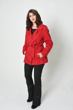 Güzel esmer kadın modelin tam boy portresi, kırmızı trençkot ceketli deri pantolon giyiyor. Duruş, kameraya doğru yürüme. Beyaz stüdyo arka planında izole.