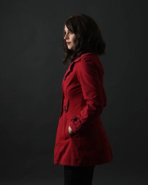 Kırmızı trençkot giyen güzel esmer kadın modelin yakın plan portresi. Karanlık stüdyo arka planında gölgelerle izole edilmiş. arka plan, kameradan uzaklaşıyor.