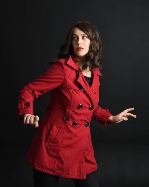 Kırmızı trençkot giyen güzel esmer kadın modelin yakın plan portresi. Karanlık stüdyo arka planında gölgelerle izole edilmiş. El hareketi uzanıyor