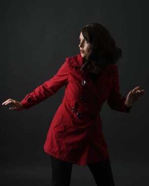 Kırmızı trençkot giyen güzel esmer kadın modelin yakın plan portresi. Karanlık stüdyo arka planında gölgelerle izole edilmiş. El hareketi uzanıyor