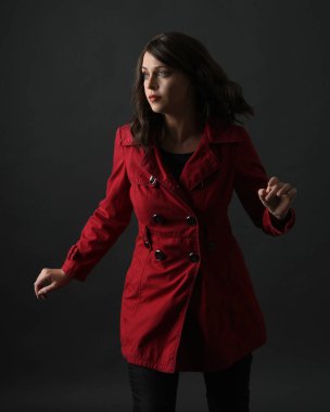 Kırmızı trençkot giyen güzel esmer kadın modelin yakın plan portresi. Karanlık stüdyo arka planında gölgelerle izole edilmiş. El hareketi uzanıyor