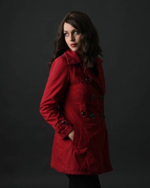 Kızıl yağmurluk giyen güzel esmer kadın modelin yakın plan portresi. Karanlık stüdyo arka planında gölgelerle izole edilmiş. Eller ceketin cebinde..